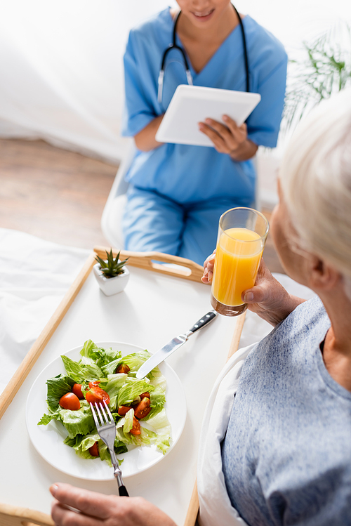 high angle view of senior woman having breakfast near nurse using digital tablet on blurred background