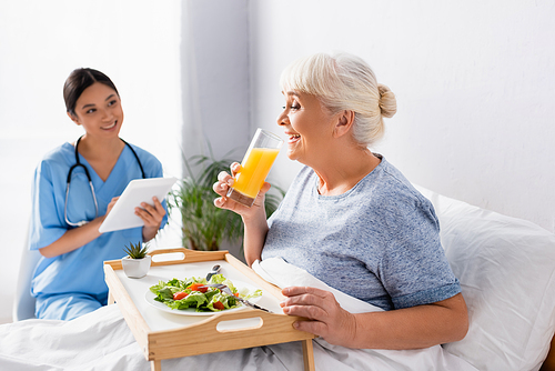 pleased aged woman having breakfast and drinking orange juice near smiling asian nurse on blurred background