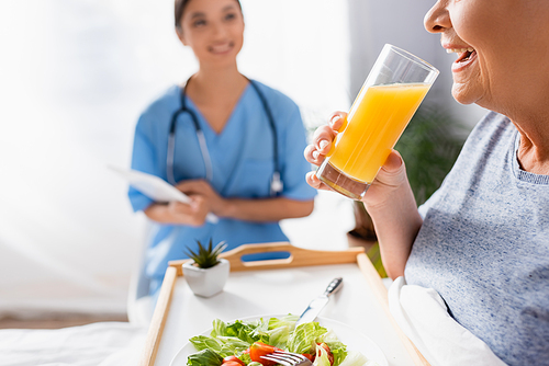 happy aged woman drinking orange juice near smiling asian nurse on blurred background