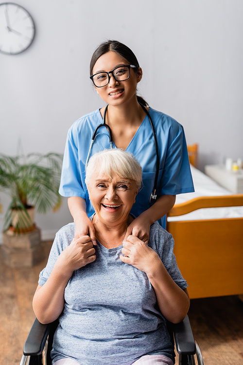 happy asian nurse and laughing elderly woman in wheelchair holding hands while