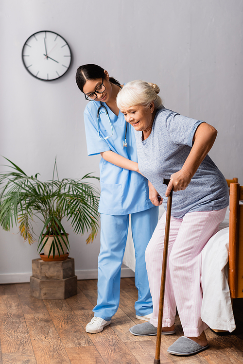 asian woman helping aged woman with walking stick getting up from bed in hospital