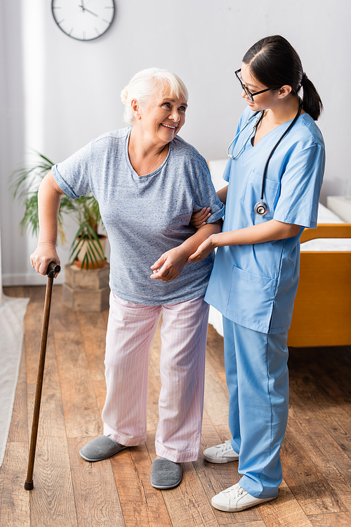 elderly woman with walking stick looking at young asian nurse supporting her in hospital