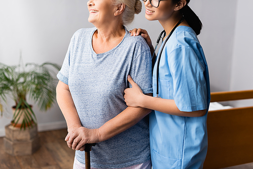 cropped view of smiling nurse supporting positive aged woman with walking stick in hospital