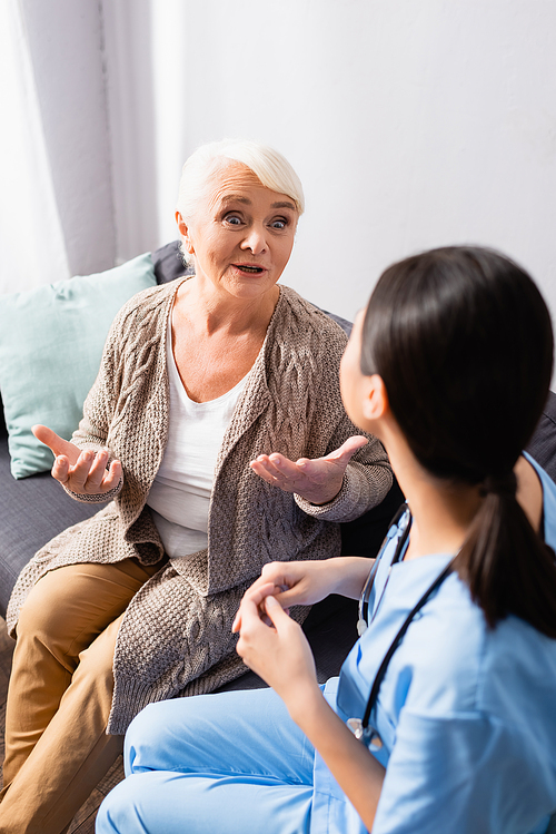 amazed senior woman gesturing during conversation with nurse in nursing home, blurred foreground