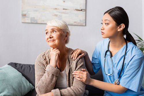 young asian nurse calming upset elderly woman sitting and looking away in nursing home