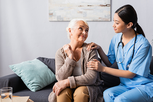 young asian nurse touching smiling senior woman during conversation in nursing home