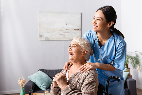 happy asian nurse touching shoulders of aged handicapped woman laughing in wheelchair