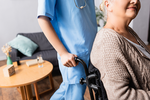 partial view of nurse near smiling handicapped woman sitting in wheelchair, blurred background