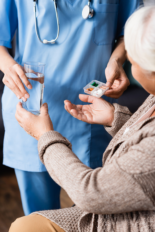 cropped view of nurse giving pills and glass of water to senior woman, blurred background