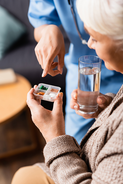 nurse pointing at pills in hand of senior woman with glass of water, blurred background