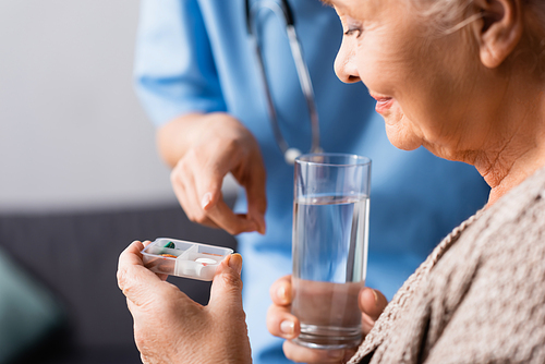 nurse pointing with finger at pills in hand of aged woman holding glass of water, blurred foreground
