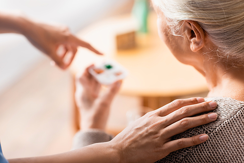 partial view of nurse touching shoulder of aged woman and pointing with finger at pills on blurred background