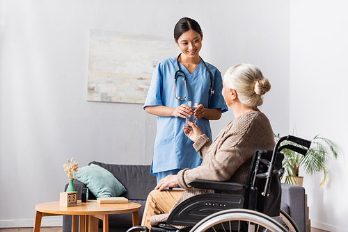 smiling asian nurse holding glass of water near aged disabled woman in wheelchair