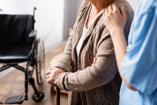 cropped view of nurse touching shoulder of elderly woman with walking stick near wheelchair on blurred background