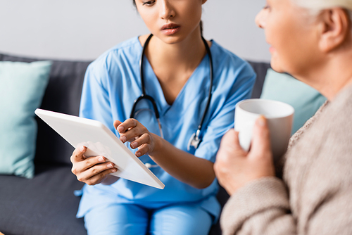 cropped view of asian nurse using digital tablet near elderly woman holding cup of tea