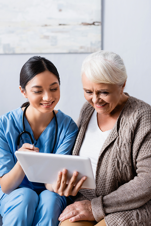 cheerful asian nurse pointing with finger at digital tablet near smiling senior woman