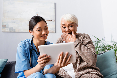 laughing senior woman covering mouth with hand while looking at digital tablet in hands of asian nurse
