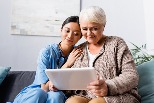 happy asian woman leaning on shoulder of aged smiling woman holding digital tablet
