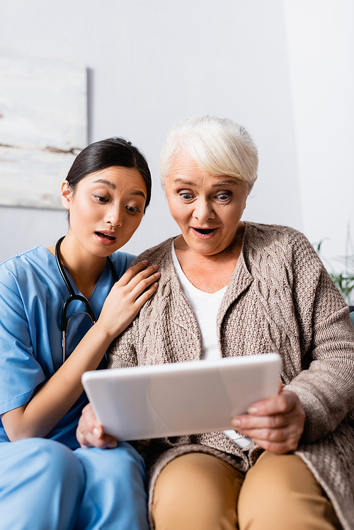 surprised asian woman touching shoulder of amazed senior woman looking at digital tablet