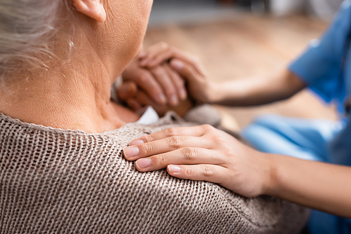 cropped view of nurse touching hand and shoulder of elderly patient in nursing home, blurred background