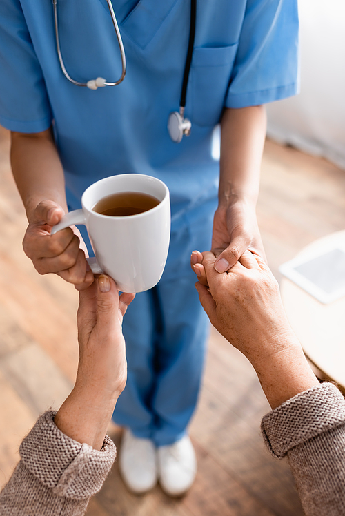 cropped view of nurse giving cup of tea to senior woman while holding her hand, blurred background