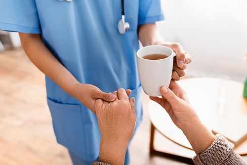 partial view of nurse holding hand of aged woman while giving her cup of tea, blurred background