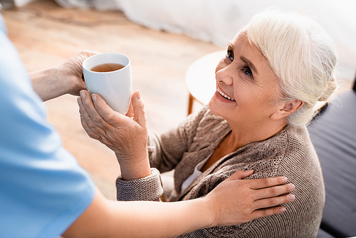 smiling senior woman taking cup of tea from nurse on blurred foreground