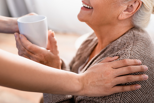cropped view of nurse touching shoulder of smiling elderly woman and giving hear cup of tea, blurred background