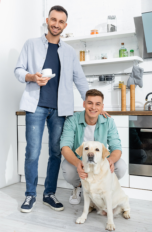 happy man holding cup and standing near husband and dog in kitchen