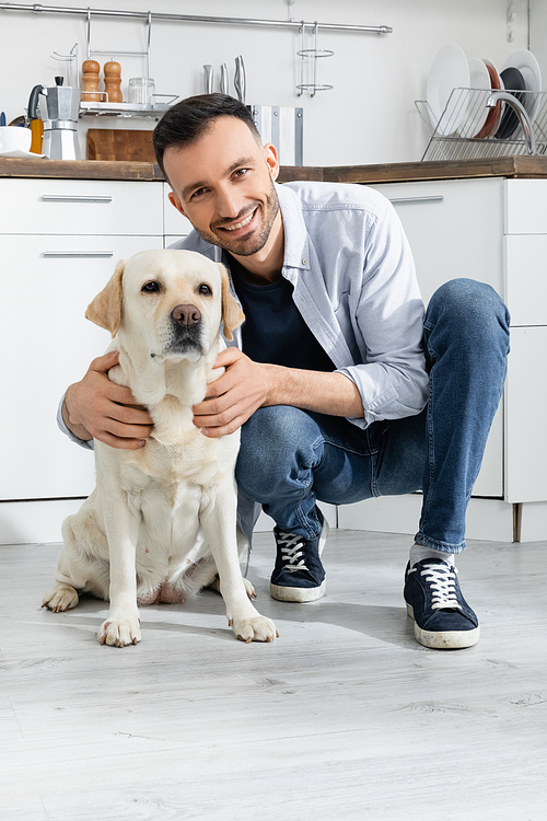 cheerful man in jeans sitting and cuddling labrador at home