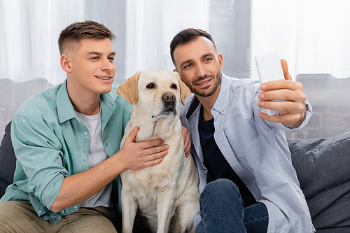 cheerful same sex couple smiling and taking selfie with labrador
