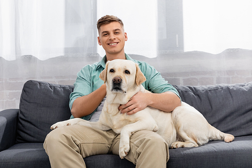 cheerful man sitting on couch and cuddling labrador