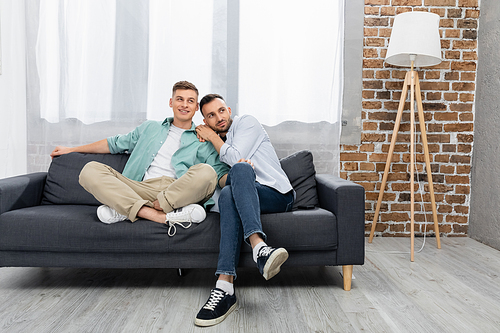 cheerful same sex couple looking away while sitting on couch at home