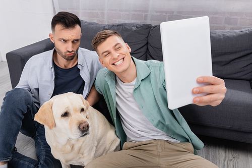 happy same sex couple taking selfie on digital tablet near labrador in living room