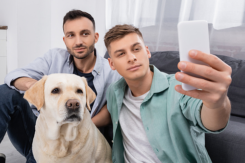 same sex couple taking selfie on smartphone near labrador in living room