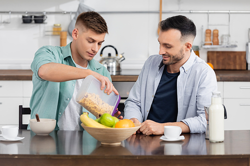 homosexual man pouring corn flakes near husband, cups and fruits