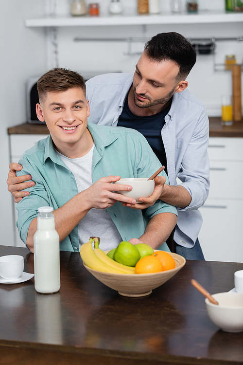 homosexual man holding bowl near happy husband and fruits on table