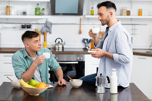 happy same sex couple looking at each other while holding cups in kitchen