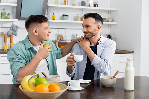 happy homosexual man holding cup and looking at husband in kitchen