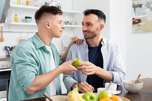 happy homosexual man giving apple to husband in kitchen