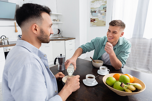 happy homosexual man pouring milk in bowl with corn flakes near husband