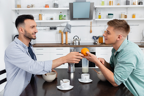 side view of happy homosexual man giving orange to husband during breakfast