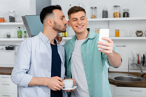 happy man pouting lips while kissing cheek of smiling husband taking selfie at home