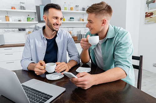 happy homosexual couple holding coffee near laptop on table
