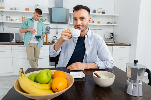 happy homosexual man drinking coffee near husband on blurred background