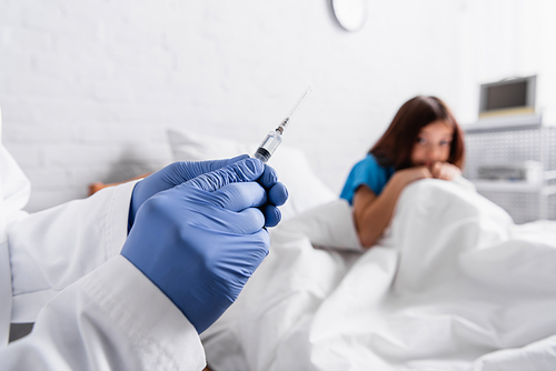 afraid child sitting on bed near doctor with syringe on blurred foreground