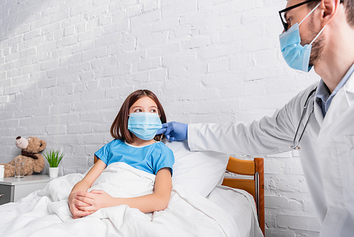 pediatrician in medical mask touching neck of sick girl sitting in hospital bed