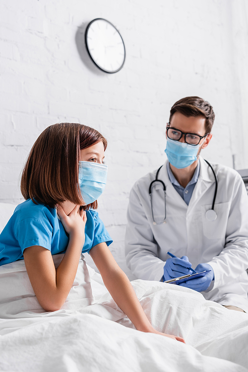 girl in medical mask touching throat near doctor writing diagnosis on clipboard