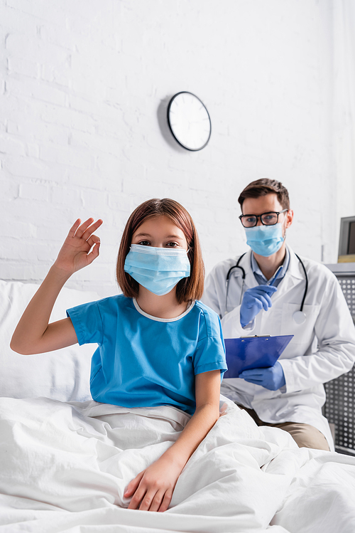 child in medical mask showing okay gesture near doctor with clipboard on blurred background