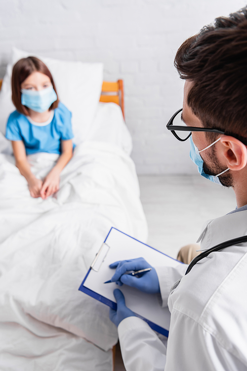 pediatrician writing diagnosis on clipboard near sick girl in medical mask on blurred background